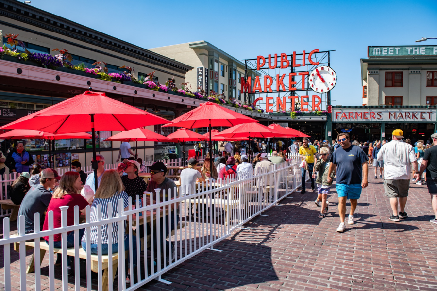 Crowds explore Seattle’s Pike Place Market under bright red umbrellas and the iconic “Public Market Center” neon sign.