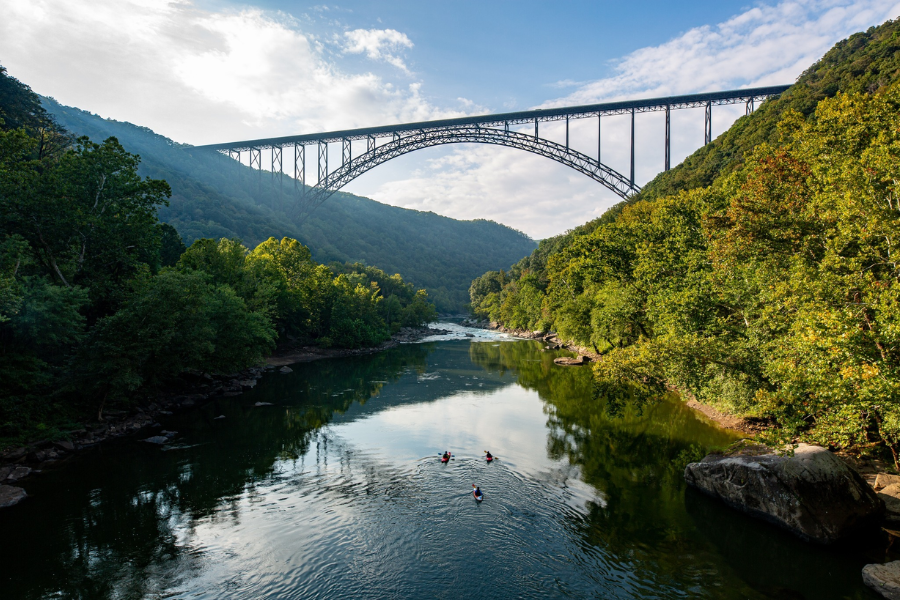 The New River Gorge Bridge stretches across a deep, forested canyon in West Virginia as kayakers paddle the calm river below.