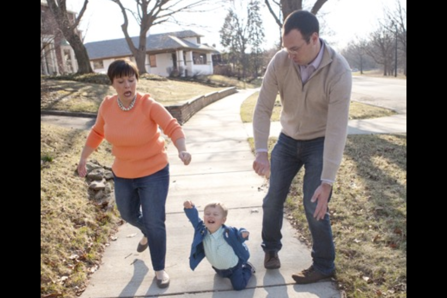 A young child falls dramatically on a sidewalk while both parents reach out in surprise, captured just too late to stop the tumble.