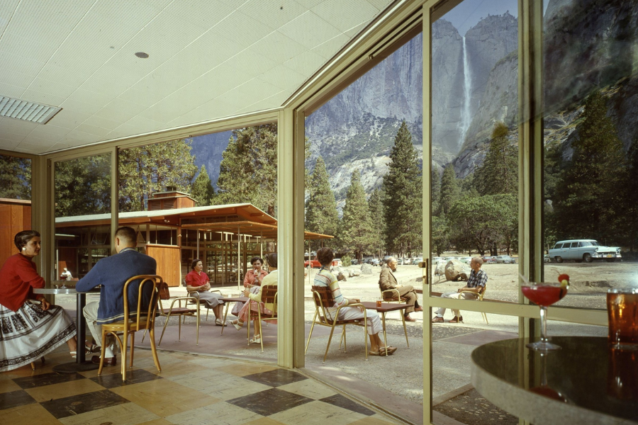 People sit both indoors and outdoors at a mid-century café with large windows offering a direct view of Yosemite Falls and towering cliffs.