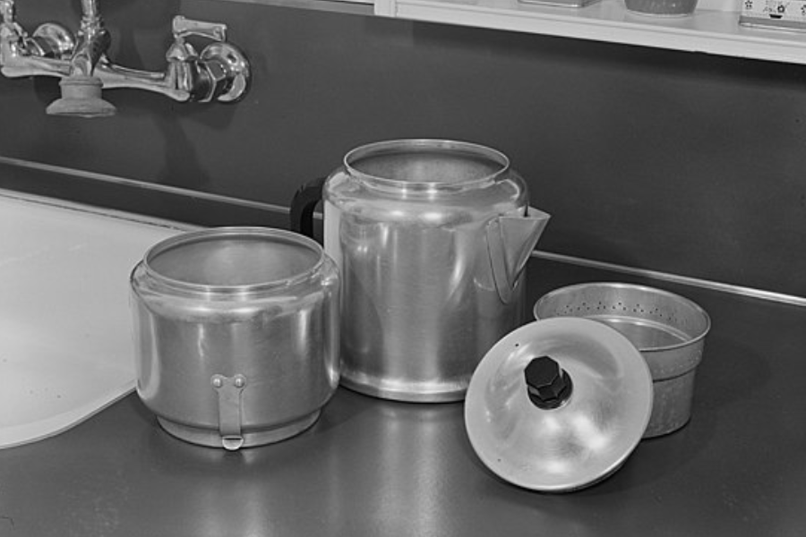 Vintage kitchen setup with metal cooking utensils including a percolator-style coffee pot, strainer, pot, and lid placed near a sink.