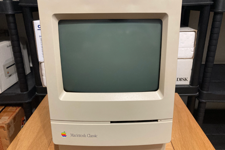 Close-up of a vintage Macintosh Classic computer with a blank screen, sitting on a wooden table in a room with boxed electronics.