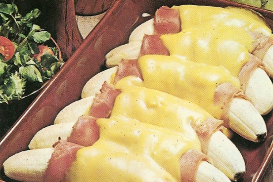 A casserole dish lined with peeled bananas wrapped in ham and topped with melted yellow cheese, served next to a leafy green salad.