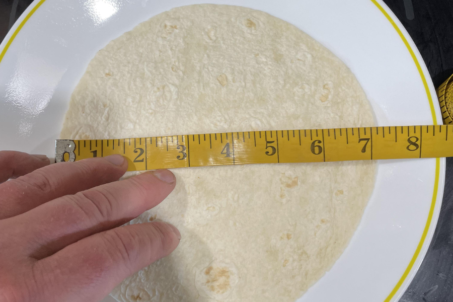 A soft tortilla measured with a yellow tape measure across its diameter, indicating about 8 inches, held on a white plate with a yellow rim.