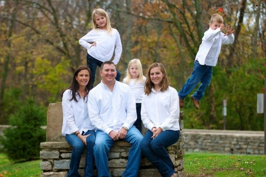 A family in matching white shirts and jeans poses outdoors, while a young boy unexpectedly jumps off the stone bench mid-shot.