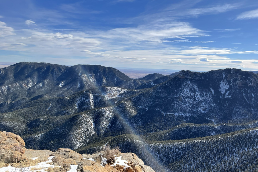 A clear winter view from a high mountain overlook shows snow-dusted pine-covered peaks under a bright blue sky with wispy clouds.