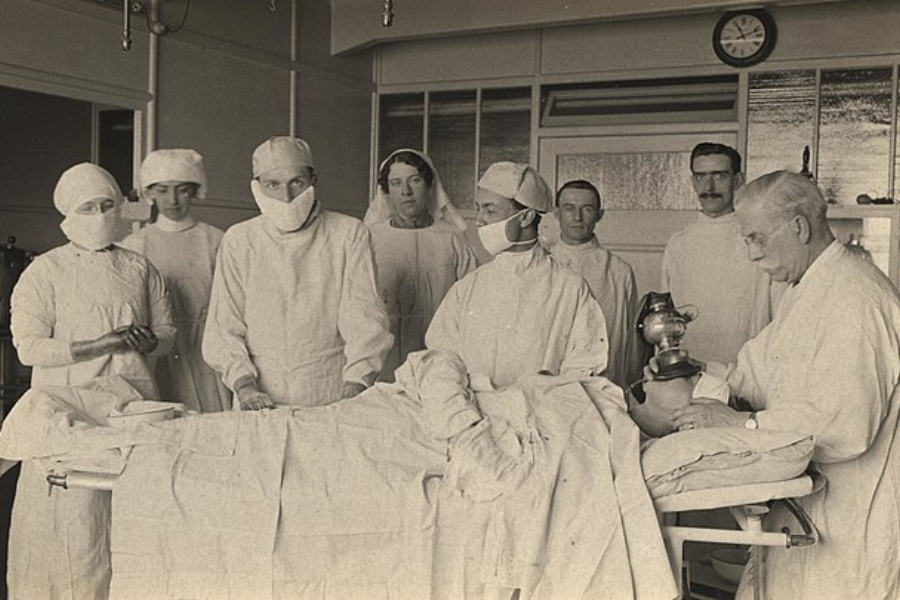 Early 20th-century surgical team in full gowns and masks preparing a patient for surgery, with an anesthetist holding a vintage ether mask.