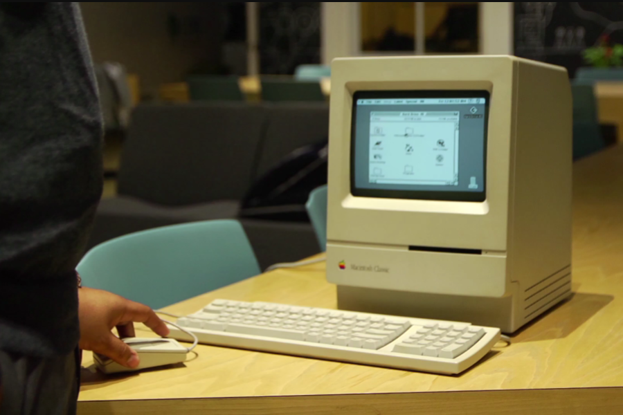 Alt Text: Person using a powered-on Macintosh Classic computer, with a hand on the mouse and a keyboard in front, set in a modern indoor space.