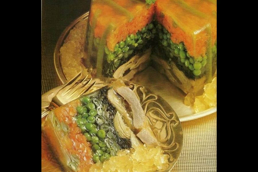 A molded gelatin dish with layers of carrots, peas, spinach, and meat slices set in clear aspic, served on a silver platter with decorative gelatin chunks.