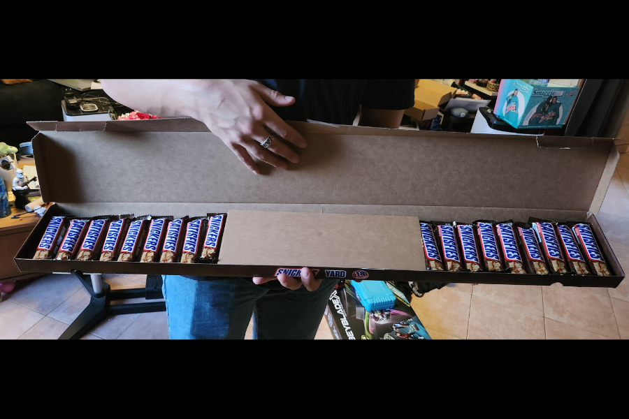 A person holding a long open box labeled “Snickers Yard” containing 18 full-size Snickers bars arranged in two rows.