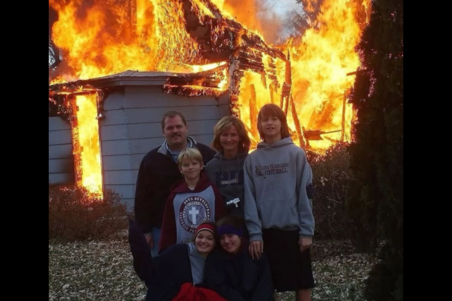 A family smiles in front of a house engulfed in flames, posing calmly as the inferno blazes behind them.