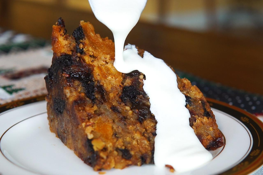Slice of plum pudding filled with dried fruits, topped with a stream of sweet white sauce being poured.