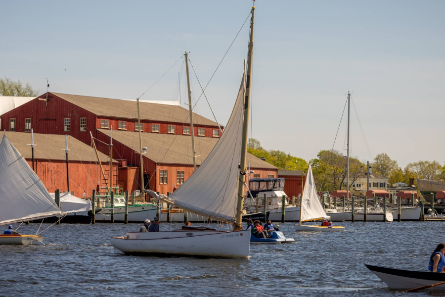 Sailboats glide across the water in front of a coastal harbor lined with rustic red buildings, with people aboard enjoying a sunny day.