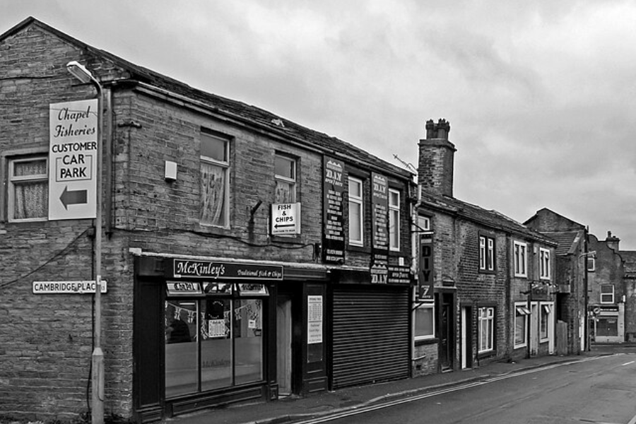 Black and white photo of a street corner in a small English town, featuring a fish and chips shop called McKinley’s and signs for Chapel Fisheries.