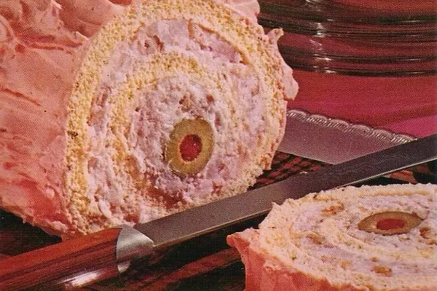 A pink frosted rolled sandwich loaf with a savory filling of meat and cream cheese, centered with a green olive with pimento, sliced like a cake roll.