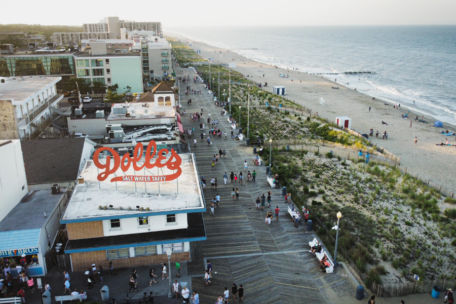An aerial view of the Rehoboth Beach boardwalk shows crowds walking past the Dolles Salt Water Taffy shop and sandy beachfront.