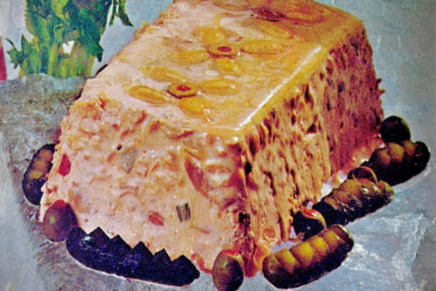 A molded meat and vegetable loaf encased in a shiny gelatin layer, decorated with slices of green olives and surrounded by pickles.