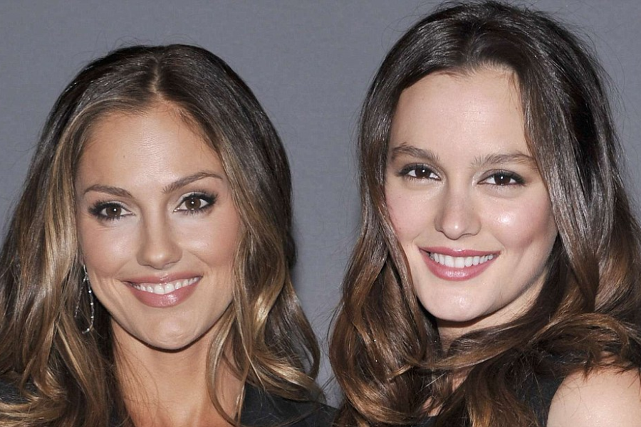 Close-up of two women with long, wavy brown hair standing side by side and smiling at the camera. Both wear subtle makeup and dark clothing against a plain gray background.