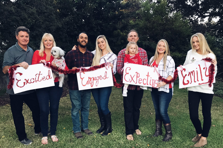 A family holds signs reading “Excited,” “Engaged,” “Expecting,” and “Emily,” each announcing a milestone, while smiling cheerfully in a backyard.