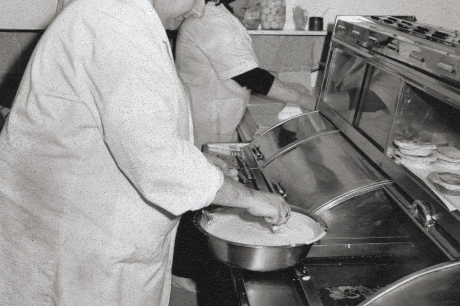 wo workers in white coats preparing food in a fish and chips shop, one of them dipping food into batter beside a fryer.