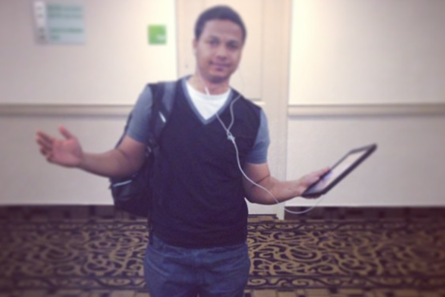 A casually dressed man standing indoors with headphones in, holding a tablet in one hand and gesturing with the other.
