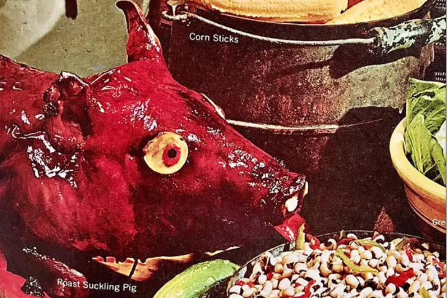A whole roasted suckling pig with glazed red skin and a dramatic apple-like eye, surrounded by side dishes including black-eyed peas and corn sticks.