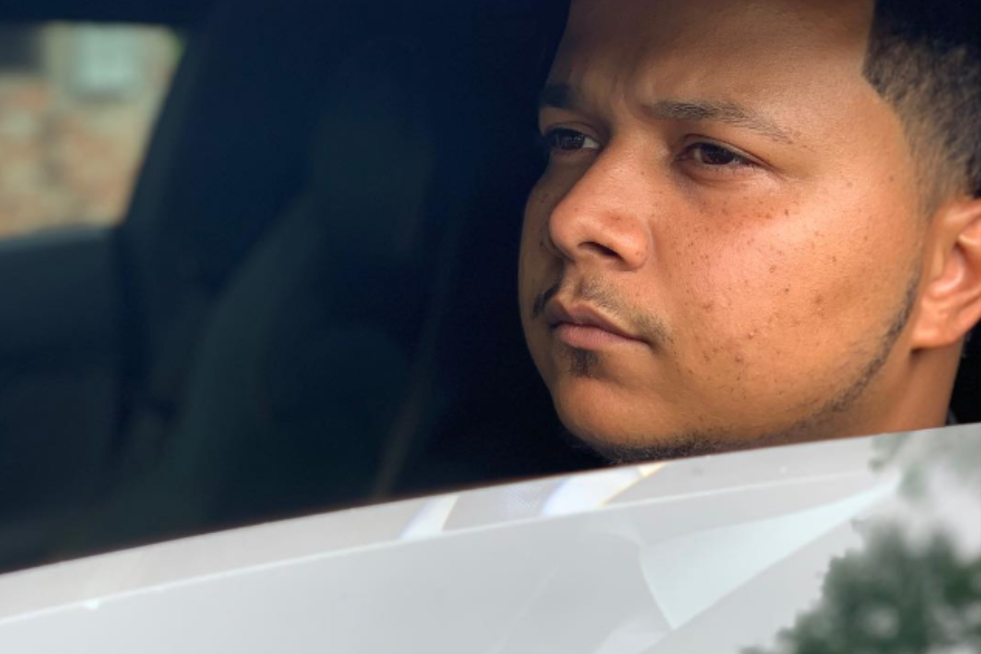 lose-up of a man with a focused expression looking out from the driver’s seat of a car, partially framed by the window.