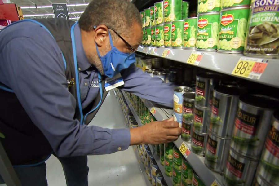 Older man wearing a blue Walmart vest and face mask arranges cans of vegetables on a grocery store shelf.
