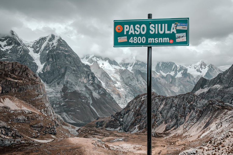 A green road sign reading "Paso Siula 4800 msnm" stands against a dramatic backdrop of steep, rocky Andean peaks dusted with snow under a cloudy sky.