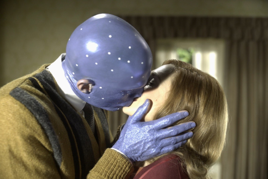 A blue-skinned humanoid with motion capture dots kisses a woman with light brown hair in a warmly lit room.