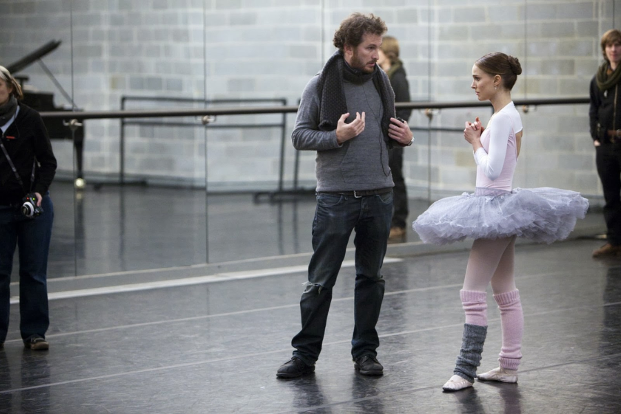 A ballet dancer in a pink and grey tutu listens intently to a director during rehearsal in a mirrored dance studio.