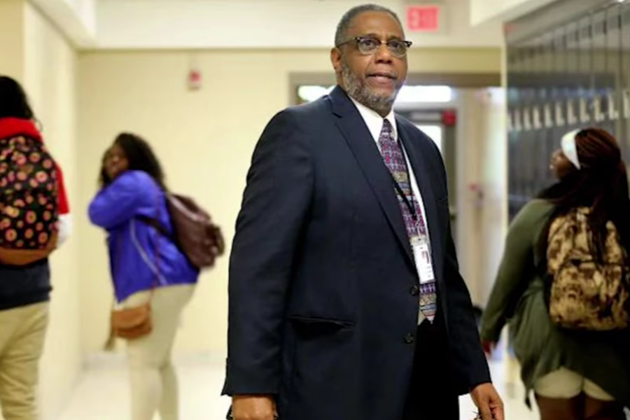 Man in a dark suit stands in a school hallway with students walking in the background.