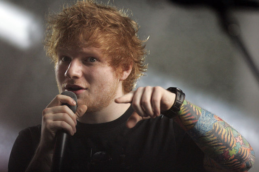 Man with tousled red hair and colorful sleeve tattoo points toward the camera while holding a microphone.