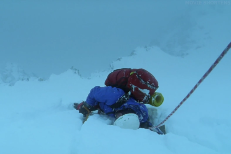 A climber lies collapsed in snow during a whiteout, roped in and partially buried with gear on their back, amid a harsh, foggy environment.