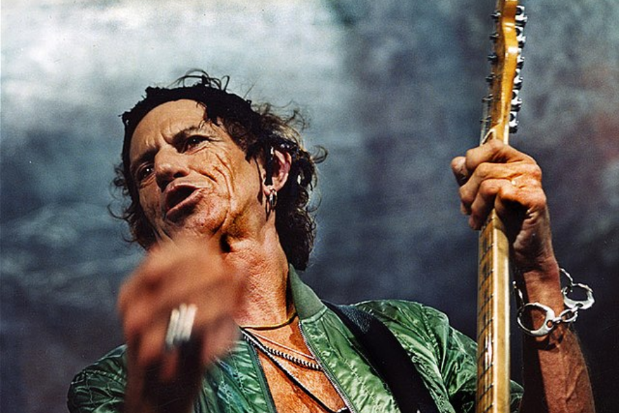 Man with weathered features and layered jewelry plays electric guitar on stage while singing with intensity.