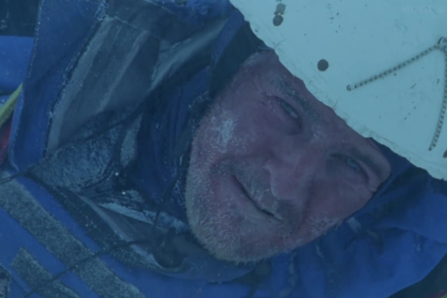 A close-up of a climber’s frost-covered face, wearing a white helmet and blue jacket, showing visible exhaustion and exposure to extreme cold.