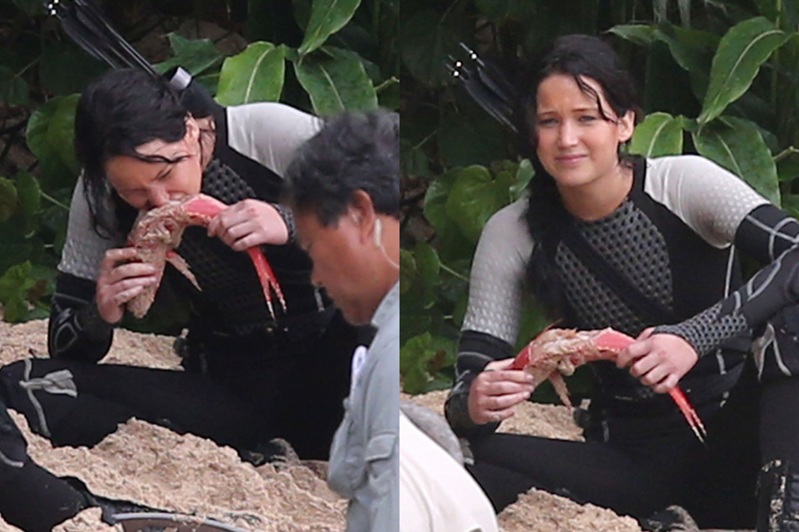 A woman in a wetsuit holds and bites into a large fish while sitting on a sandy beach during a film shoot.