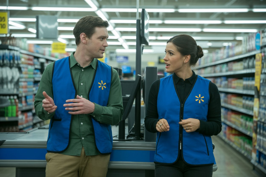 Male and female Walmart employees in blue vests talk at a checkout counter in the store aisle.