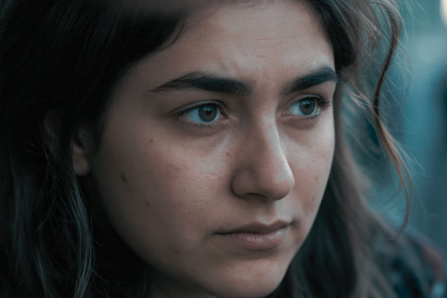 A close-up of a teenage girl with messy hair and a solemn, distant expression. Her face is softly lit, revealing a mood of sadness or introspection.