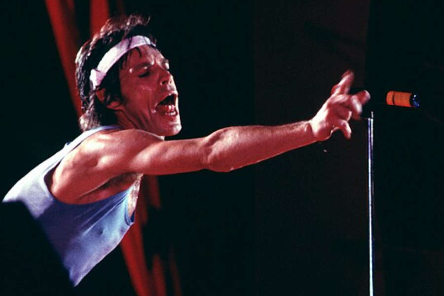 Man in a blue tank top and headband passionately sings into a microphone on stage with an outstretched arm.