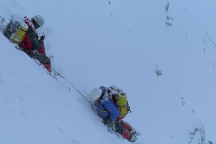 Two climbers roped together make a slow ascent up a steep snow-covered slope, each carrying yellow oxygen tanks and ice axes.