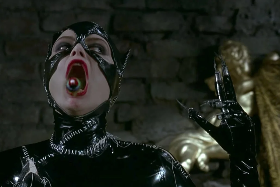 A woman in a stitched black leather catsuit dramatically tilts her head back with her mouth open wide, revealing a colorful item inside.