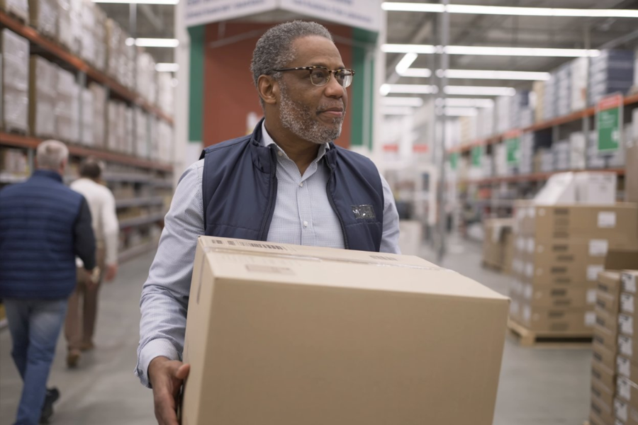 Walmart employee carries a large cardboard box through a warehouse-style store aisle lined with stacked merchandise.