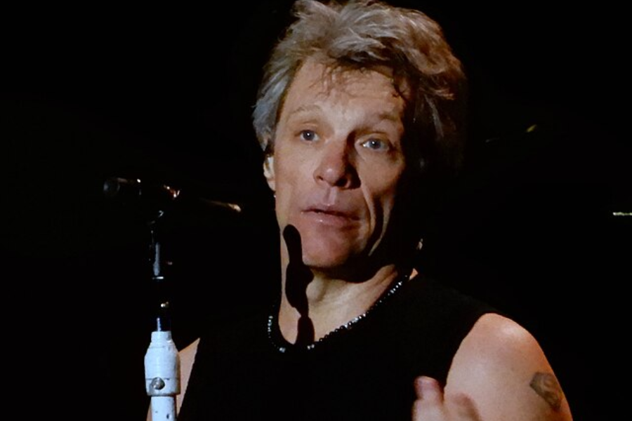 Man with light hair and a sleeveless black shirt stands at a microphone under stage lighting, mid-sentence or singing.
