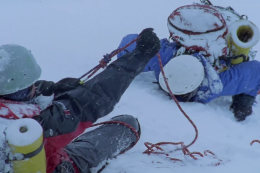 One climber in red braces the rope as another in blue struggles to stay upright in deep snow, both covered in frost and snowflakes.