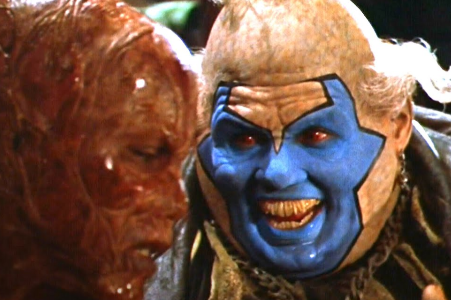A grotesque blue-faced character with sharp teeth grins menacingly next to a scarred, burned figure in a dark setting.