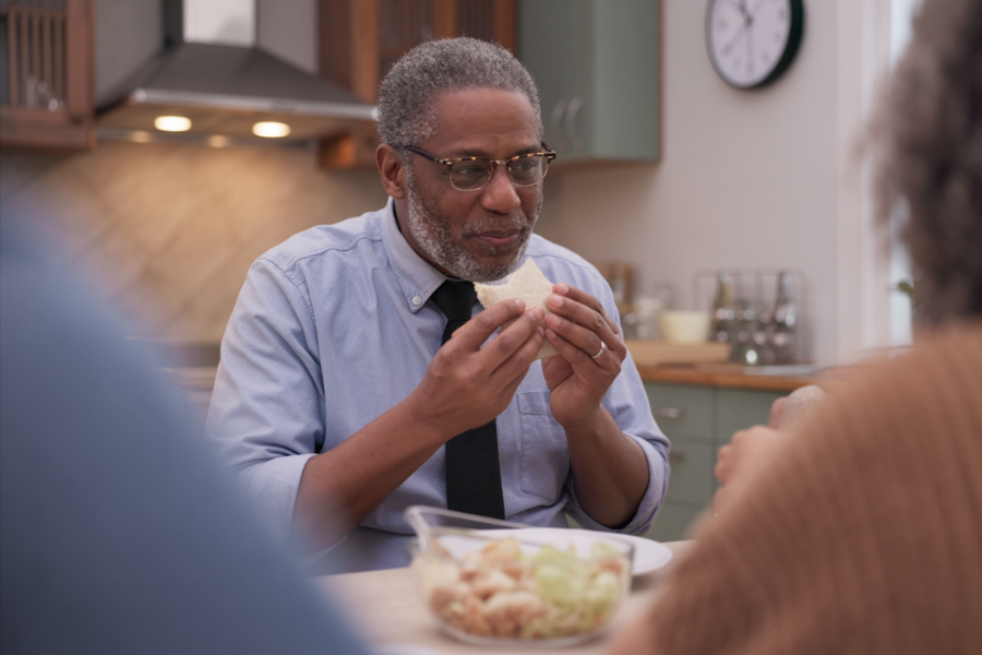 Man in a blue shirt and black tie eats a sandwich at a kitchen table with a salad bowl nearby.
