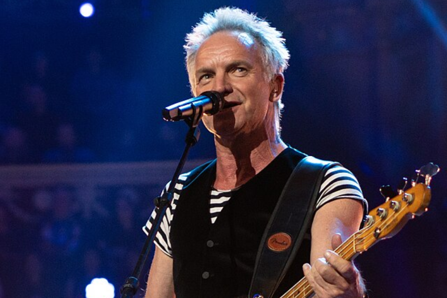 Man in a black vest and striped shirt sings and plays bass guitar under blue stage lights.