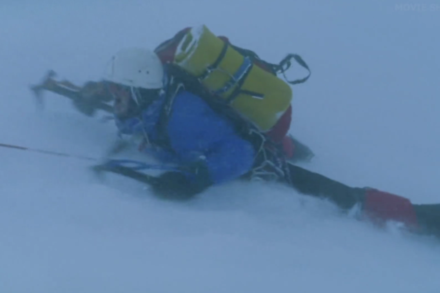 A climber in a white helmet and blue gear lies flat in the snow during a storm, gripping an ice axe and tethered by rope.