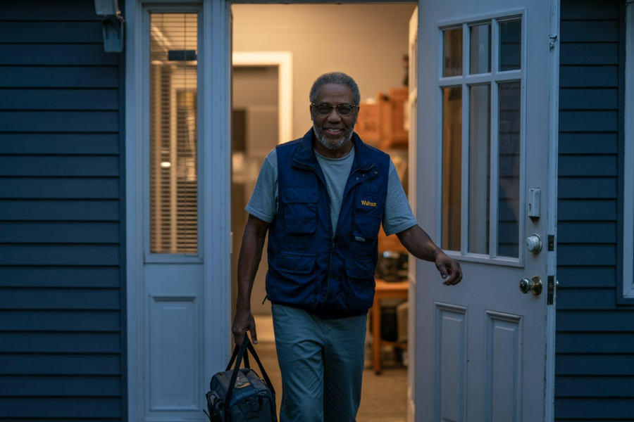 Smiling Walmart employee in a navy vest leaves home in early morning light carrying a black work bag.